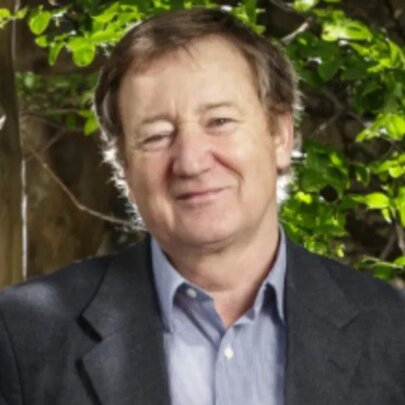 Presenter John Patrick, smiling in a garden setting, wearing a blue shirt and dark jacket.