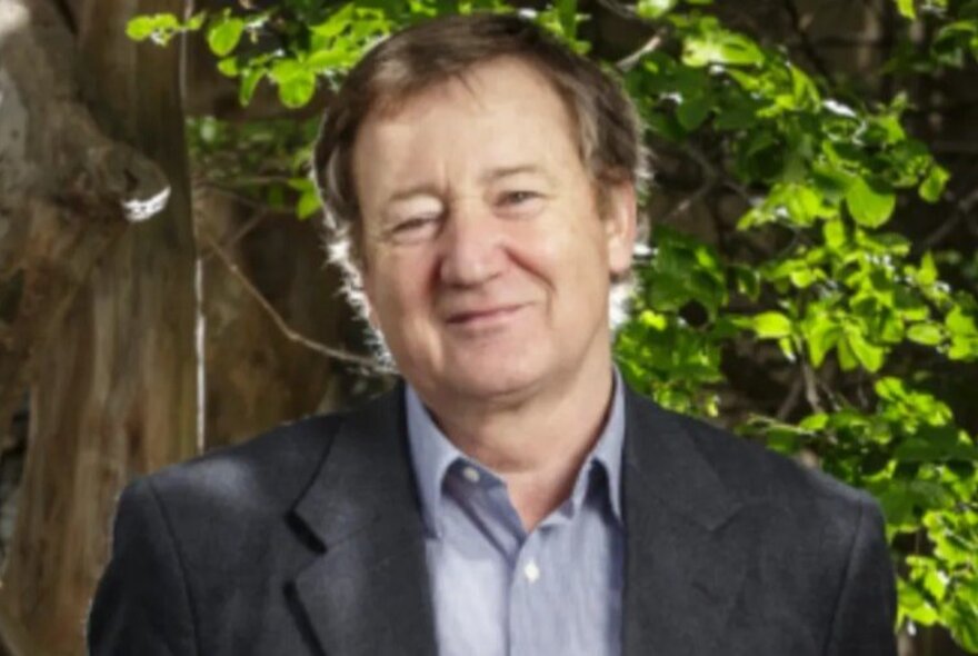 Presenter John Patrick, smiling in a garden setting, wearing a blue shirt and dark jacket.
