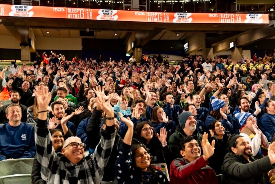 A large crowd of people in the seats at the MCG all looking upwards and smiling in warm clothing.