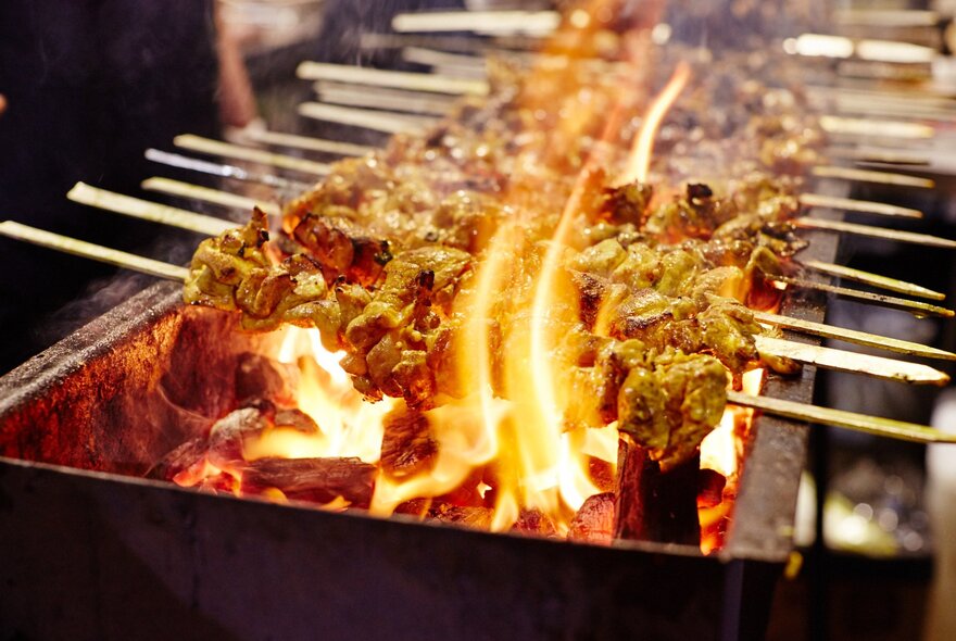 Satay chicken skewers cooking over a flame grill.