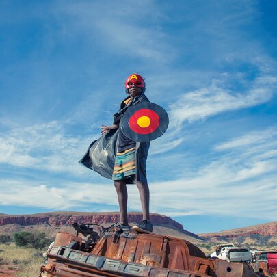 An Indigenous Australian youth stands atop a rusted, overturned car in a red desert landscape under a vast blue sky with wispy clouds. They wear a red headband and hold a large circular shield painted with concentric rings of black, red, and yellow.