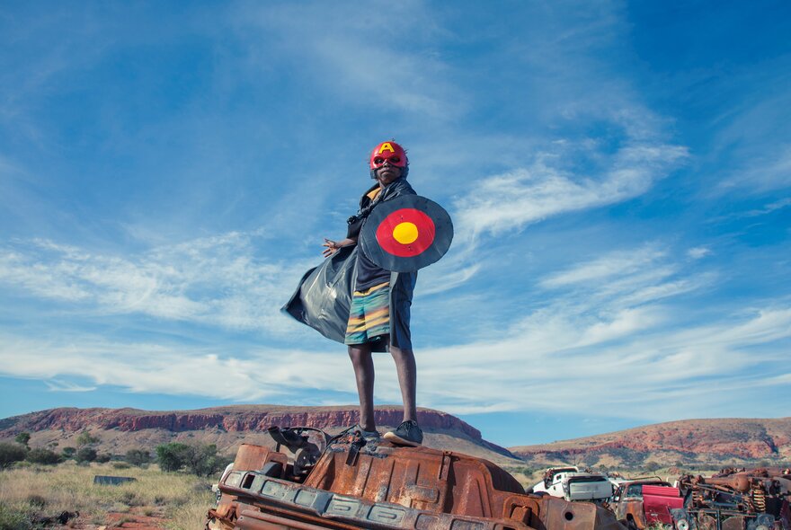 An Indigenous Australian youth stands atop a rusted, overturned car in a red desert landscape under a vast blue sky with wispy clouds. They wear a red headband and hold a large circular shield painted with concentric rings of black, red, and yellow.