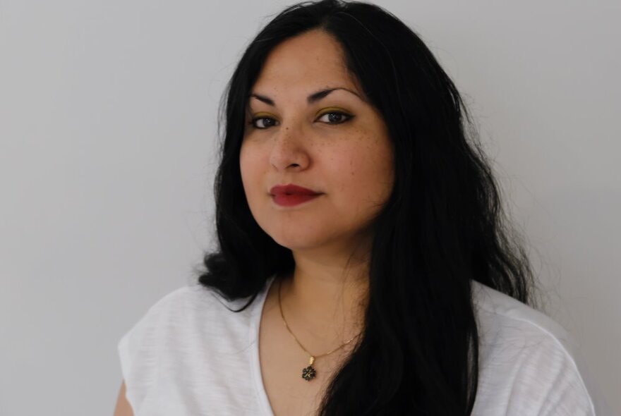 Portrait of a young woman with thick, dark hair, calmly looking at the camera in front of a white background.