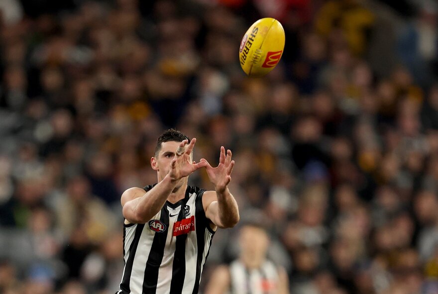 A Collingwood AFL player with his hands raised above him about to take a mark on the footy field, a blurred crowd of fans behind him.
