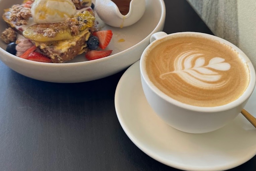 A cup of coffee next to a breakfast plate of fruit and toast.