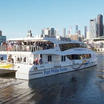 A large ferry with people on the top deck cruising on the Yarra River with the Melbourne skyline in the background. 