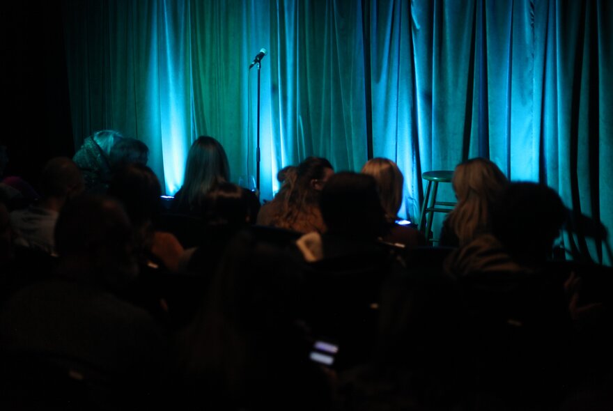 A live audience waiting for a show to begin on a empty stage shows a blue curtain and a microphone stand.