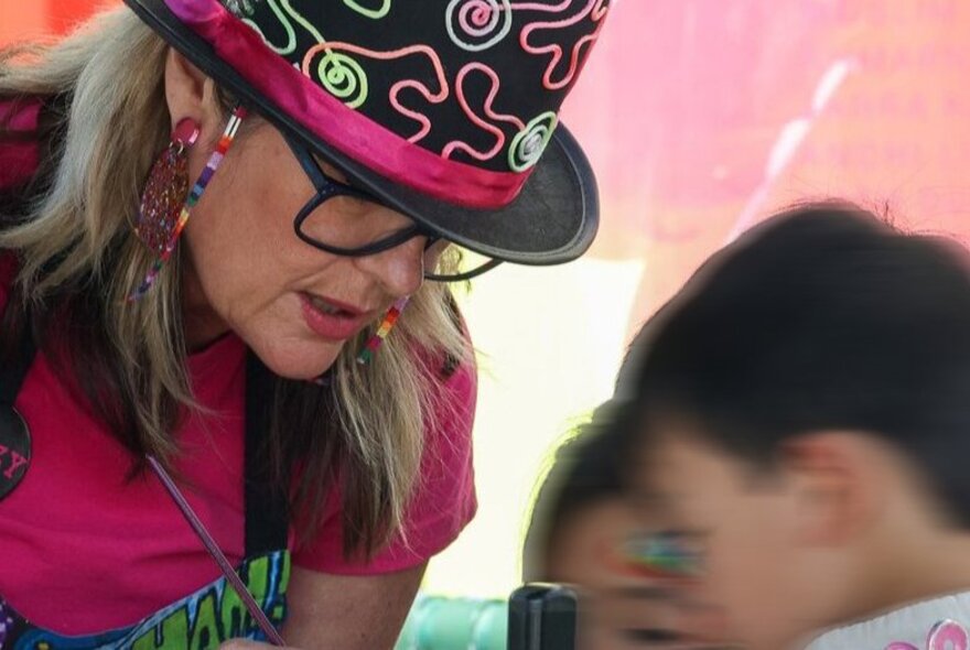 A woman wearing a bright hat and holding a thin paintbrush, painting children's faces at a street festival.