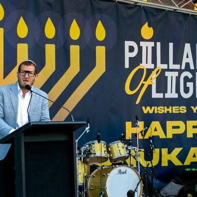 A person wearing a light blue suit jacket speaking at the Pillars of Light Chanukah celebration at Federation Square in Melbourne, he stands at a lectern in front of a banner which displays an image of a lit Menorah and the words 'Pillars of Light'. 