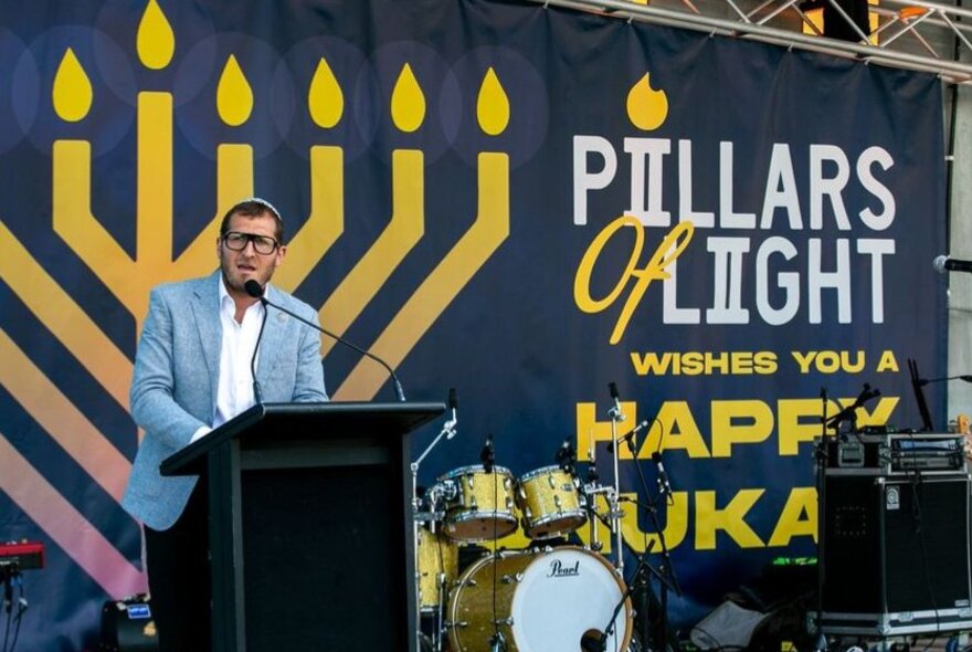 A person wearing a light blue suit jacket speaking at the Pillars of Light Chanukah celebration at Federation Square in Melbourne, he stands at a lectern in front of a banner which displays an image of a lit Menorah and the words 'Pillars of Light'. 