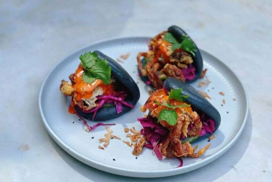 Light grey plate on white marble table. Three soft shell crab black bao buns with coriander and sauce.