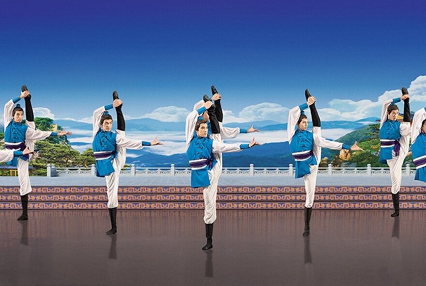 Five Chinese dancers all standing on one leg with the other leg raised high over their head on a stage against a landscape backdrop. 