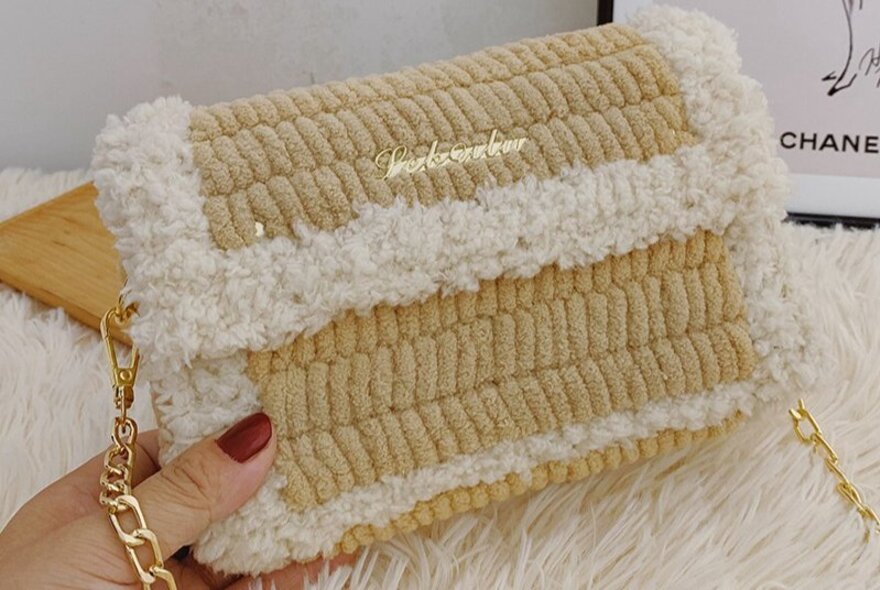 A beige and white knitted shoulder bag with plush trim and a chain strap, made in a cross-stitch workshop.