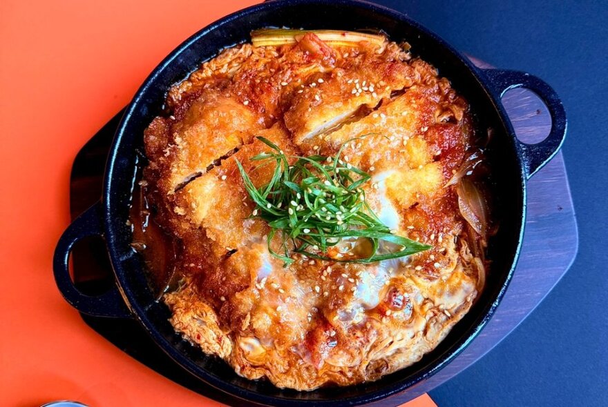 A Japanese dish cooked in a round black pot with green garnish on a table that's orange and blue