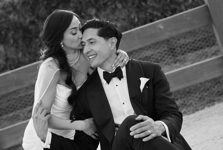 A seated groom wearing a tailored suit, with his bride kissing his head from behind.