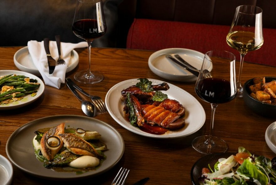 Overhead view of several dishes of food on a wooden table including meat, vegetables, cutlery, plates and wine.