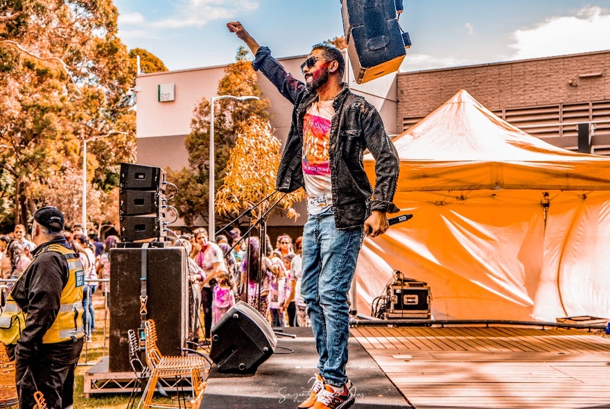 A person on an outdoor stage performing and singing at Melbourne's outdoor Holi Festival.