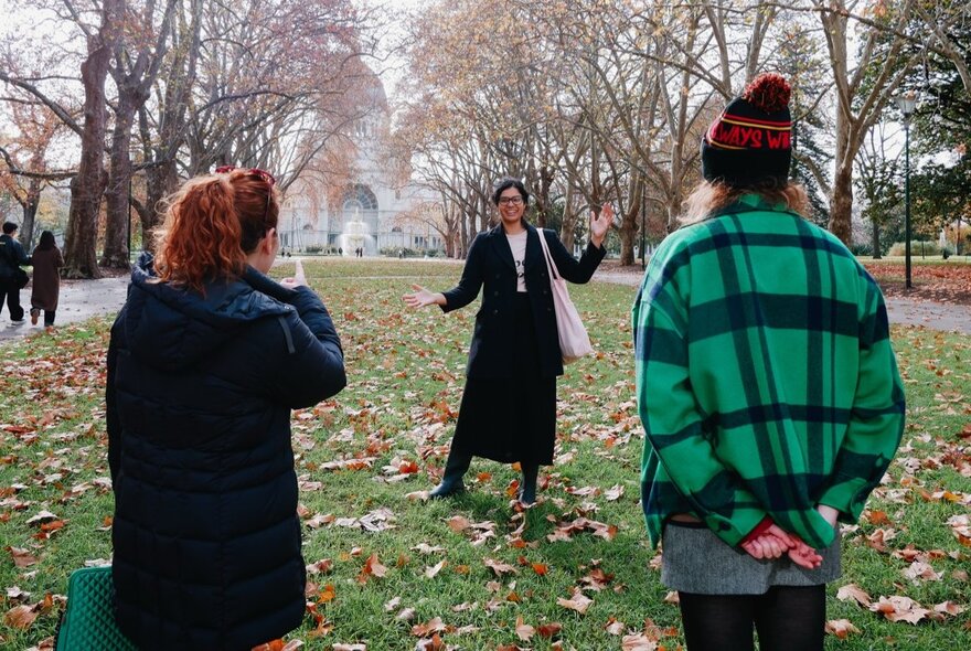 A tour guide talking to people in the Carlton Gardens, autumn leaves on the grass, the Royal Exhibition Building just visible in the background.