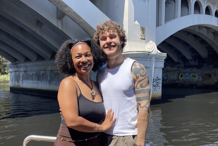 A smiling couple standing up on a sightseeing cruise boat, a view of Princess Bridge and the Yarra River behind them.
