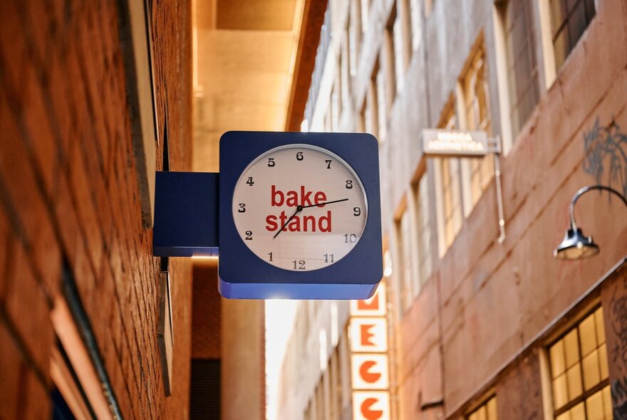 The exterior signage for the BakeStand shop, and a clock, protruding from a red brick wall in a Melbourne laneway.