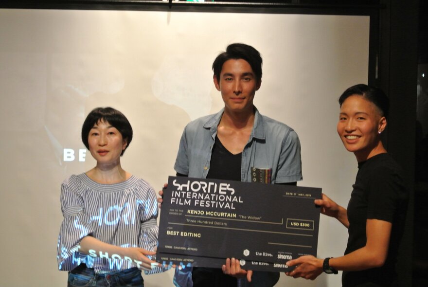 Three people in front of a projection screen, one accepting an award printed on large cardboard. 