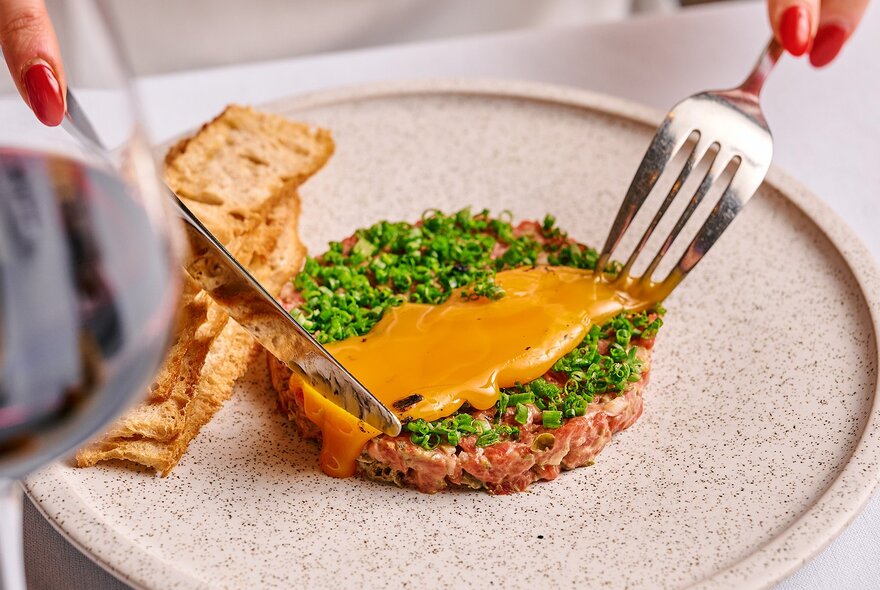 A knife and fork about to cut into a small, delicate tart with chives and an orange sauce, on a dinner plate.