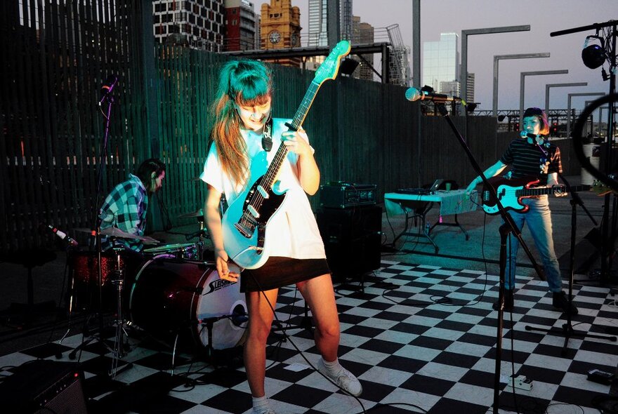 Group of young musicians performing on a rooftop with chequerboard floor, city buildings in the background.