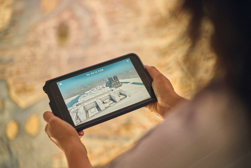 A digital device in focus, being held by two hands and showing foundations of a building, in front of a blurred, out of focus map.