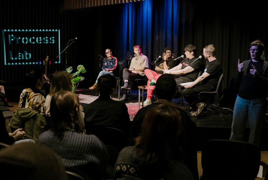 A panel of experts seated in chairs on a stage talking and engaging in front of a live audience.