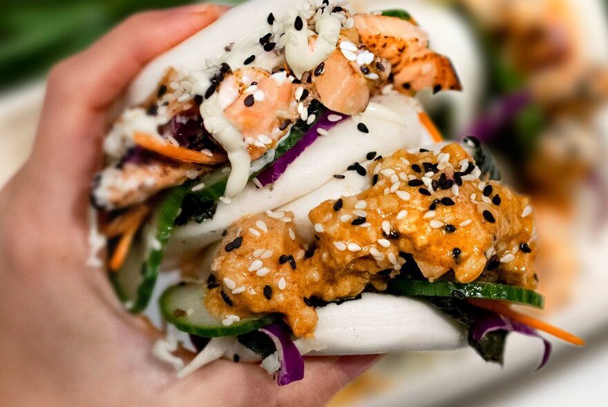 Two Japanese-inspired bao buns gripped by a hand and filled with cured salmon, cucumber and black sesame seeds.