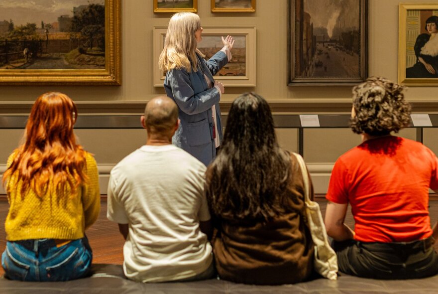 A small tour group seated while listening to a woman pointing at paintings on a gallery wall.