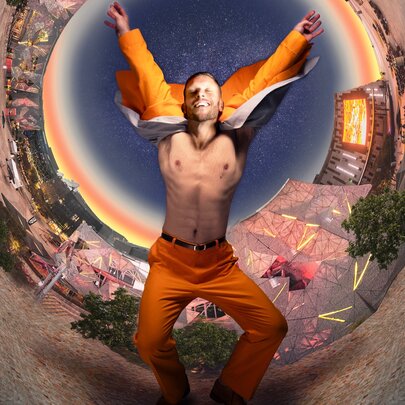 A shirtless man wearing a bright orange suit jumps enthusiastically inside a distorted, circular panoramic view of a city square under a sunset sky.