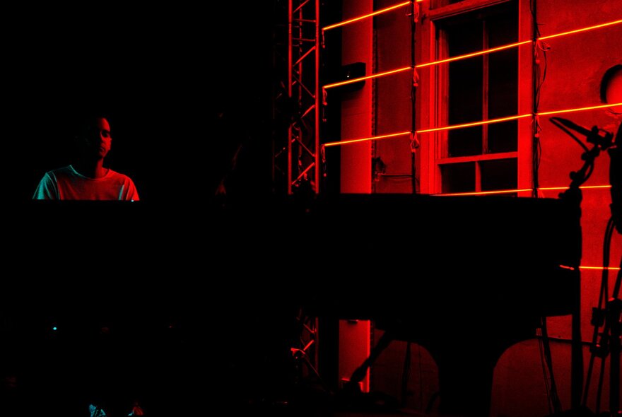 Pianist Josh Cohen playing a grand piano in a dark space lit only by red light. 
