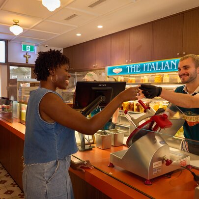 A woman smiling as she receives a taste of meat from a smiling man behind the deli counter. 