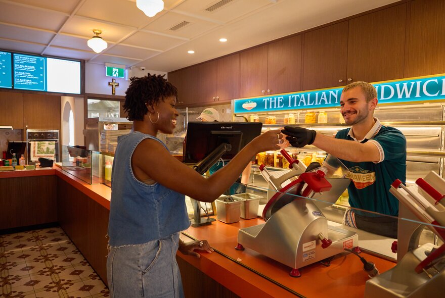A woman smiling as she receives a taste of meat from a smiling man behind the deli counter. 