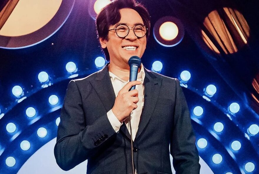 Comedian, Michael Hing, on stage in front of circular rows of bright blue lights, holding a microphone and smiling.