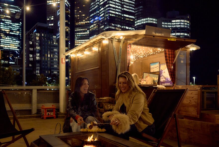 Two people roasting marshmallows over a fire pit in front of a warmly lit cabin in the city at night.
