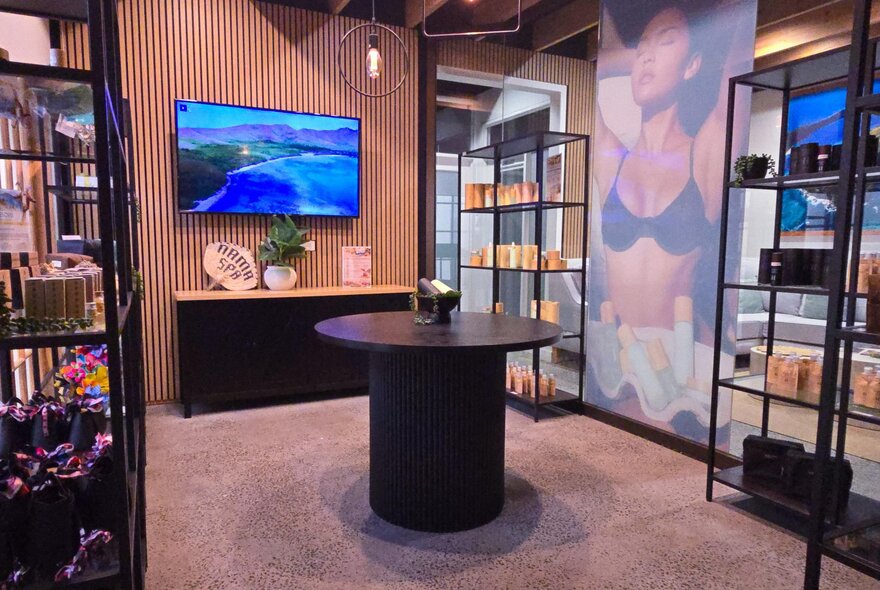 Interior of a wellness retail space featuring black metal display shelves, a central round table, wood-slat wall accents, and a large screen.