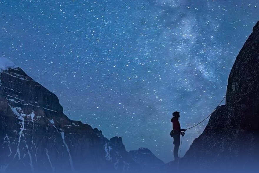 A man standing at the base of a mountain climb holding a rope, in the middle of the night with a star studded sky behind him. 