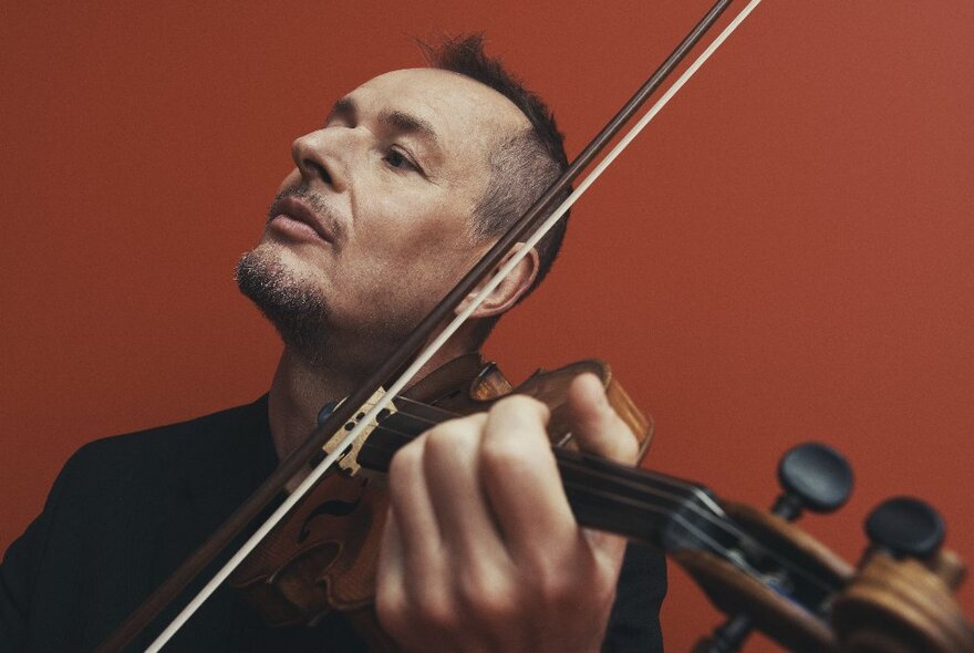 Richard Tognetti playing violin with his head tilted upwards against a russett coloured background. 