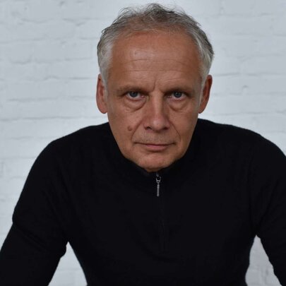 Dutch  cellist Pieter Wispelwey looking stern, framed against a white-painted brick wall wearing black.