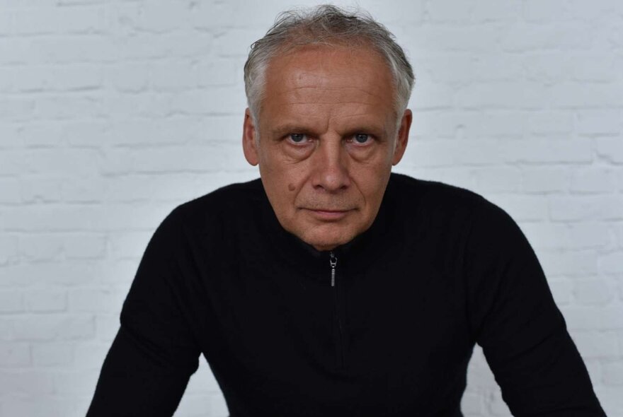 Dutch  cellist Pieter Wispelwey looking stern, framed against a white-painted brick wall wearing black.