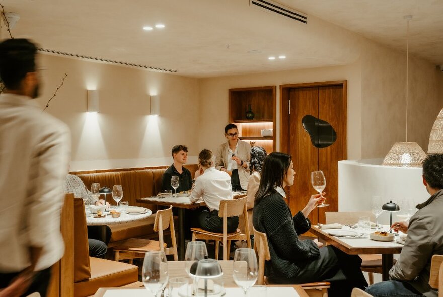 Interior of a softly lit restaurant with people dining and drinking at tables, and waiting staff walking around. 