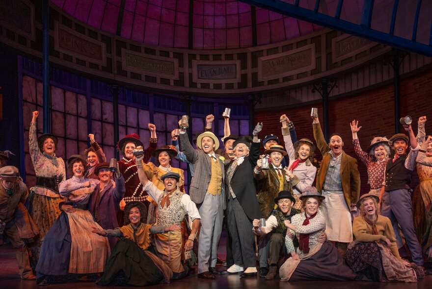 A scene from the musical My Fair Lady, with a large ensemble cast on stage, most raising a hand in the air