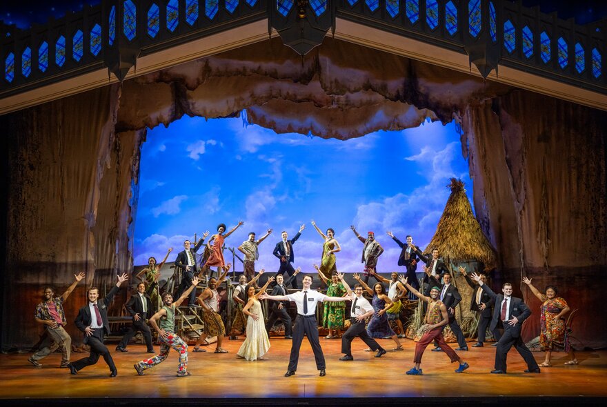The full cast of the musical Book of Mormon on stage singing and dancing, with a painted sky coloured background and other theatre props.