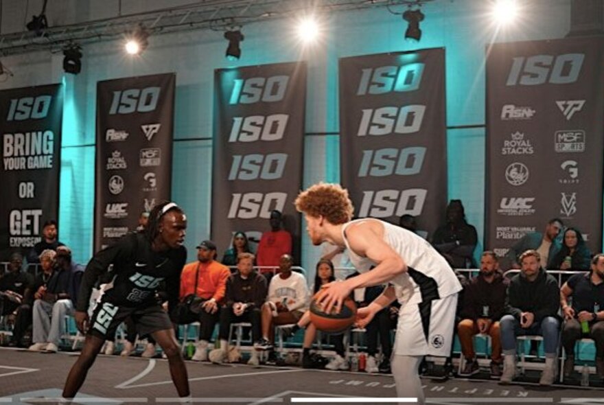 Two 1v1 basketball competitors on court, one wearing white and the other wearing black; people watching from the sideline.