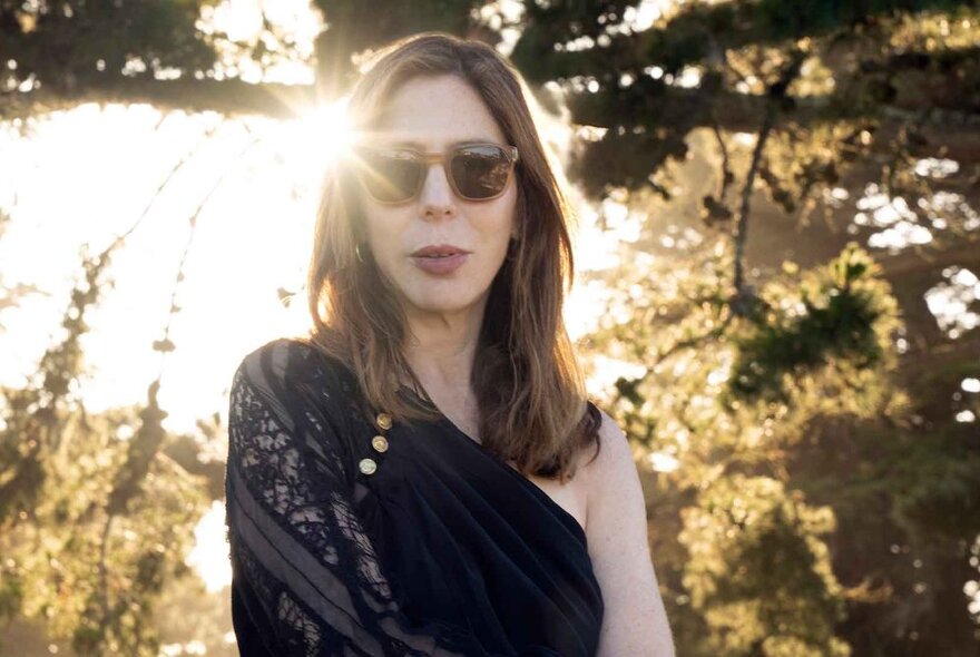 Author wearing off-the-shoulder black lace top and sunglasses, standing in front of trees with sunlight streaming through.