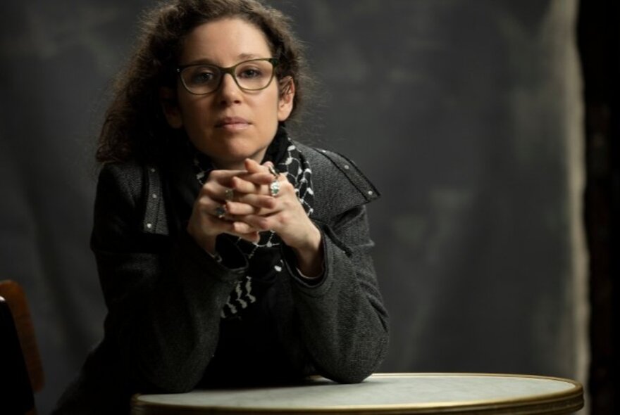 Micaela Sahhar, a woman with dark brown hair, wearing glasses, with her hands clasped in front of her, leaning forward onto a table.