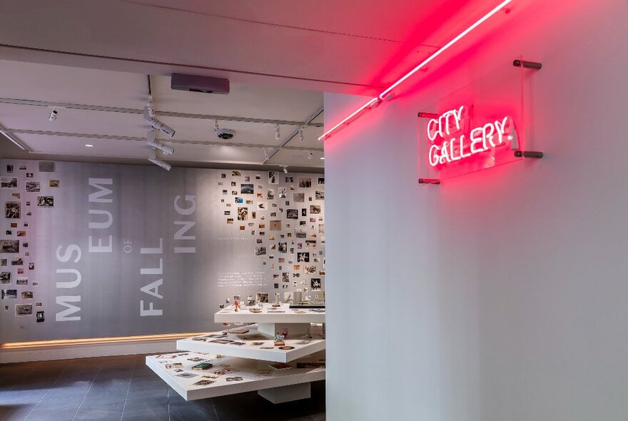 Installation view of an exhibition at City Gallery, with a pink neon sign on the wall reading CITY GALLERY and a display of images on  the rear wall. 