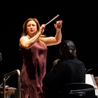 Concert conductor Simone Young AM, conducting on stage in a plum-coloured dress, musicians around her.